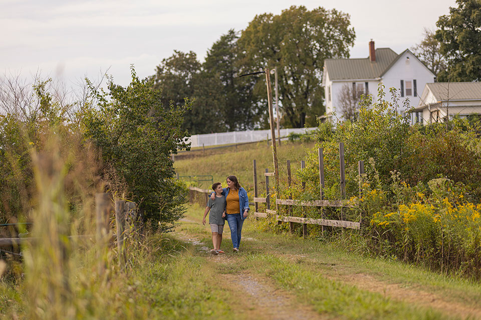 A woman and child walk arm in arm down a grassy gravel path with a house in the distance.