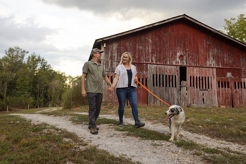 A man and woman walk a dog on a leash past an old barn.